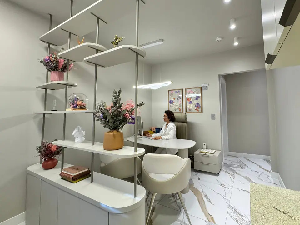 Consultório com estante de flores e a Dra. Kennya Henriqueta à mesa, ao fundo